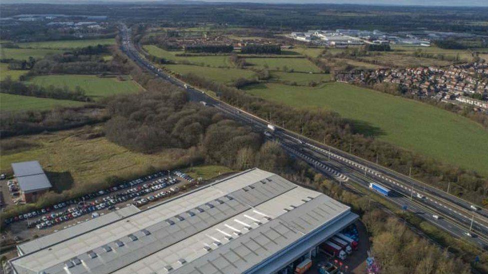 Proposed link road with fields and woods on either side. On the lefthand side of the carriageway there is a warehouse building and a car park with most of the spaces occupied and on the other side of the carriageway there is a residential development and more industrial buildings. There are chevrons and road markings on the link road.