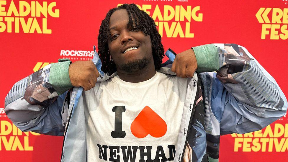 A man wearing a white t-shirt and blue patterned overcoat grabbing the top of his shoulder. The background is a red boarding with "Reading Festival" written in yellow.