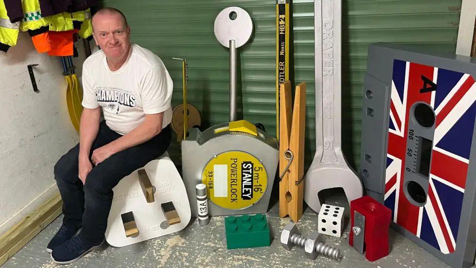a man sitting on top of a big plug with other big objects near him such ass a peg, a tape measure, a pencil sharpener and a cassette