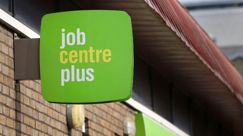 A green job centre plus sign on a brick outdoor wall 