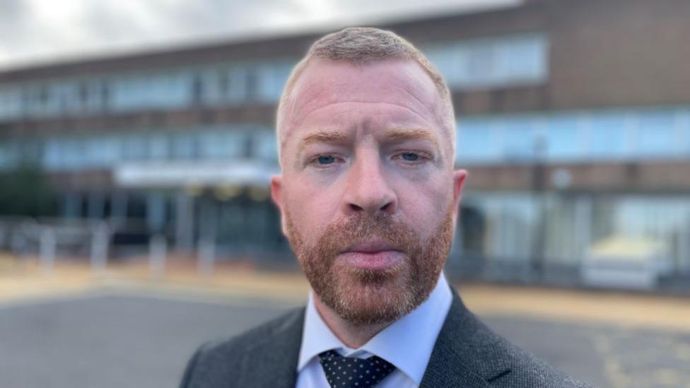 DCI Wright is wearing a grey suit and tie. He has short ginger hair and a cropped ginger beard. He is standing in front of Police Scotland headquarters at Fettes in Edinburgh.