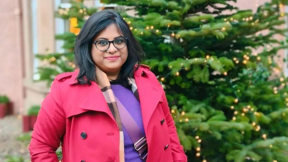 A woman with dark shoulder-length hair and glasses in a bright pink coat stands next to a Christmas tree and smiles at the camera.