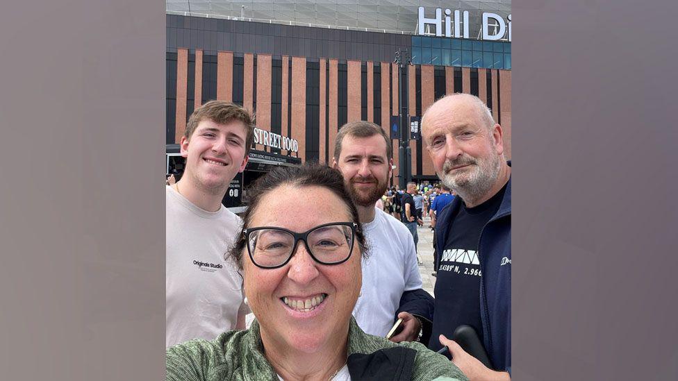 Cathy Clarke has dark hair tied back and is wearing dark-rimmed glasses. She is taking a selfie and is at the front of the photo with her husband, son and nephew standing behind.