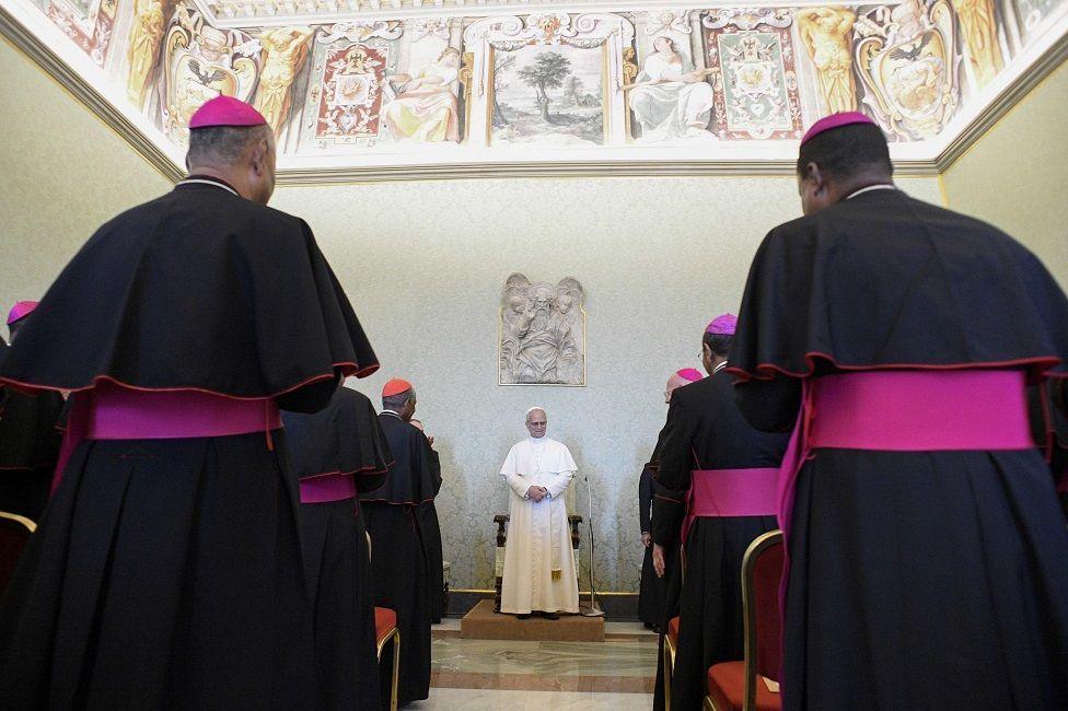 Bishops dressed in black and purple cassock rise from their seats, as the Pope stands at the front of the room and addresses them.