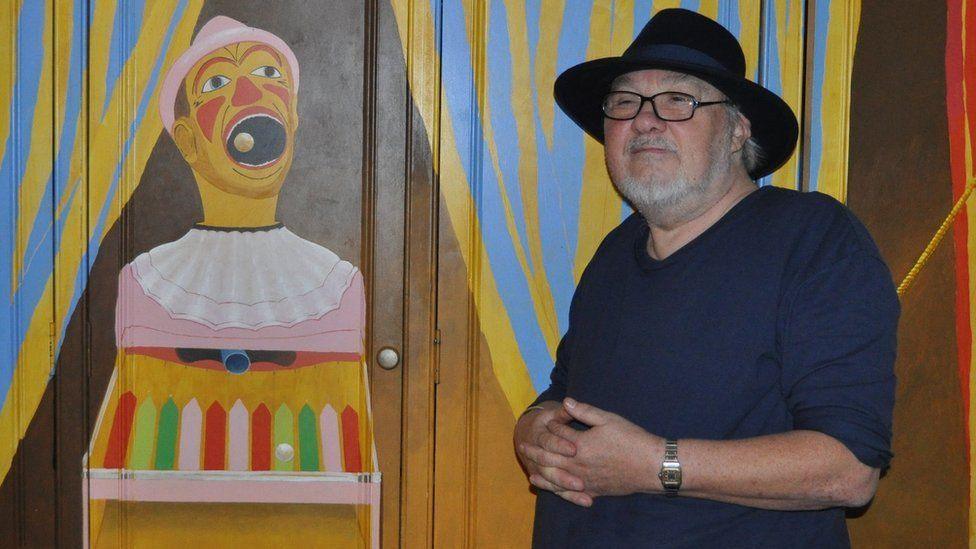 Pete Sinfield wearing fedora-type hat, standing in front of a wardrobe with a painted front of a fairground clown game where balls are fired into its mouth