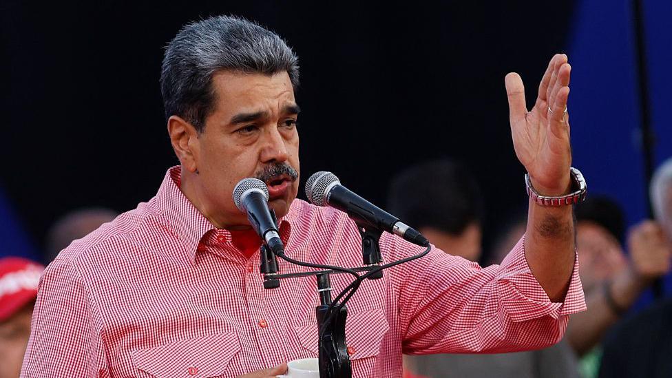 Venezuelan President Nicolás Maduro wears a red shirt and speaks into a microphone. He is gesturing with his hand.