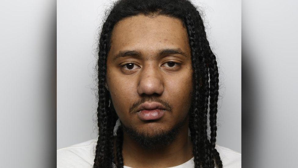 Custody picture of Kyran George. He is wearing a white t-shirt and has long dark braids.