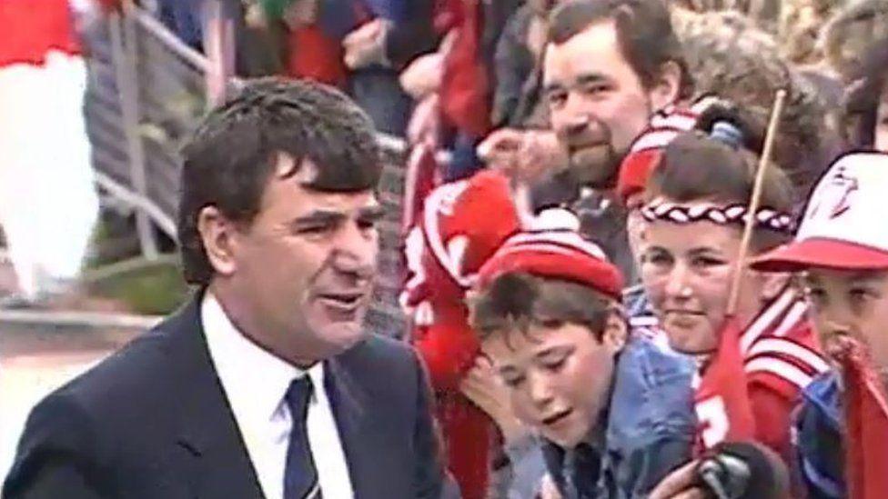 Manager of the 1989 Derry City side Jim McLaughlin walks past a packed terrace of fans dressed in the clubs colours of  red and white