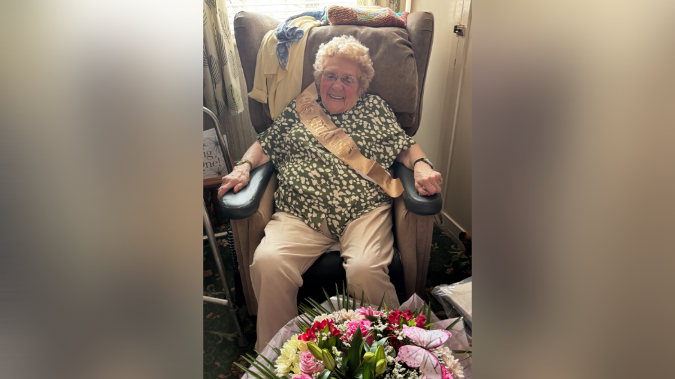 Phyliss Marshall sat inside her home with a big bouquet of flowers in front of her. She is wearing a green blouse with white spots across it. She is smiling and has a birthday sash across her.