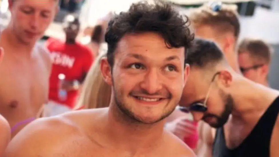 George Low, bare-chested with dark hair and stubble, smiles as he looks towards the sky, surrounded by other similarly-dressed holidaymakers 