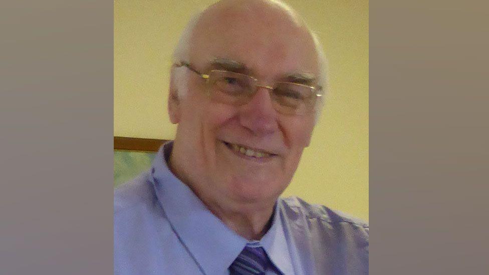 Peter Jackson who has very short white hair, is wearing metal-framed glasses and smiling at the camera. He is wearing a light blue shirt with a blue striped tie.