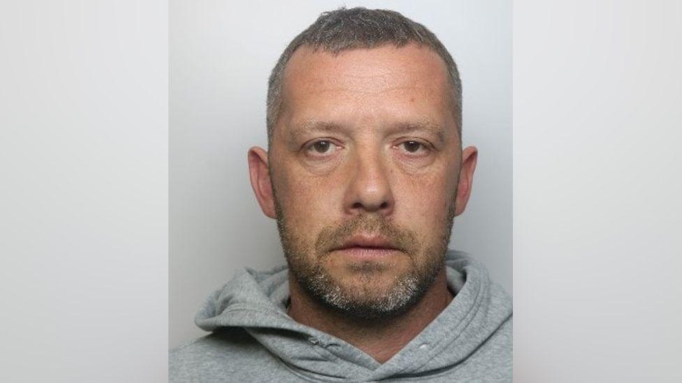 A custody shot of Kevin Newson. He is wearing a grey hoodie and is staring into the camera. He has short brown hair, flecked with grey, a short beard of the same colour.