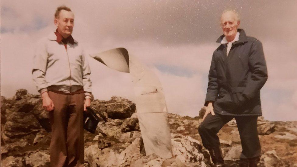 A faded colour photo of James Bolton and Robert Dalton in 1982 at the newly erected memorial to Wing Cdr Surplice. James is wearing a beige windcheater and brown trousers and Robert is wearing a grey thigh length coat over black trousers. Between them is a twisted piece of aeroplane propeller imbedded in rocks.