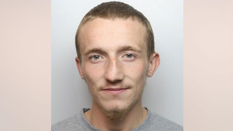 A mugshot of a man with a short brown hair, beard and moustache. He is wearing a grey top and is looking straight into the camera.