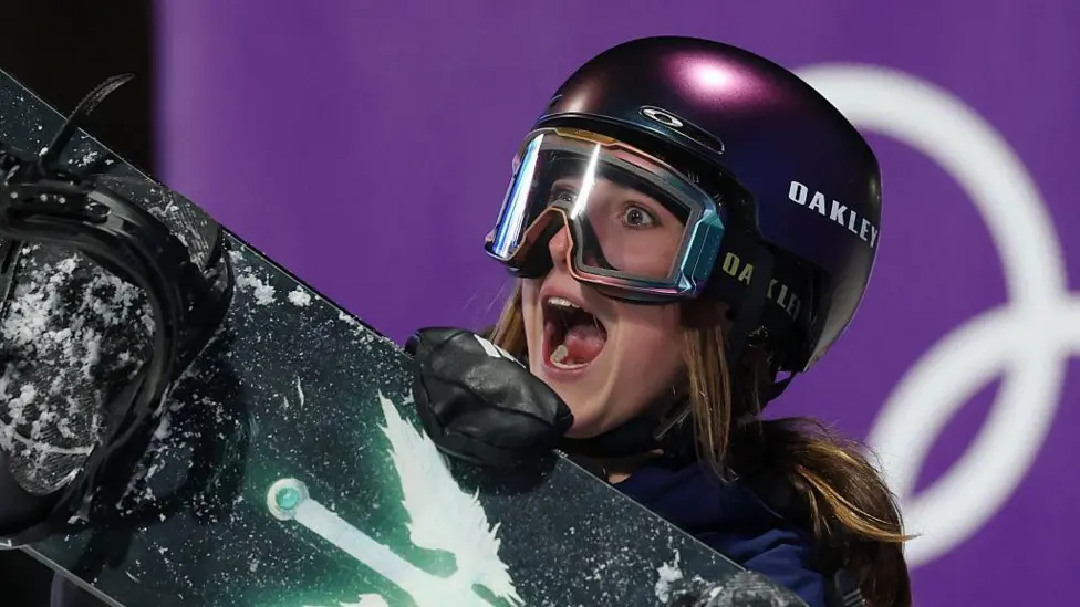 Mia Brookes shows an expression of delight, with her mouth wide open as she wears a protective dark-coloured helmet and ski goggles while carrying a snowboard