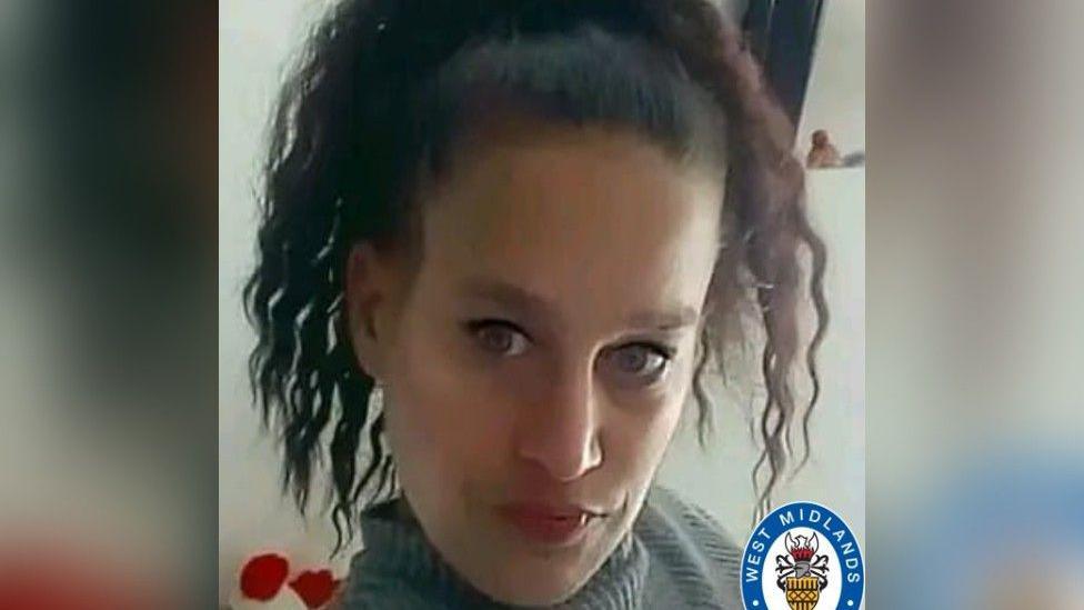 A woman with curly dark hair and black eyeliner, wearing a grey turtleneck, looks at the camera. The West Midlands Police logo is at the bottom right of the picture.