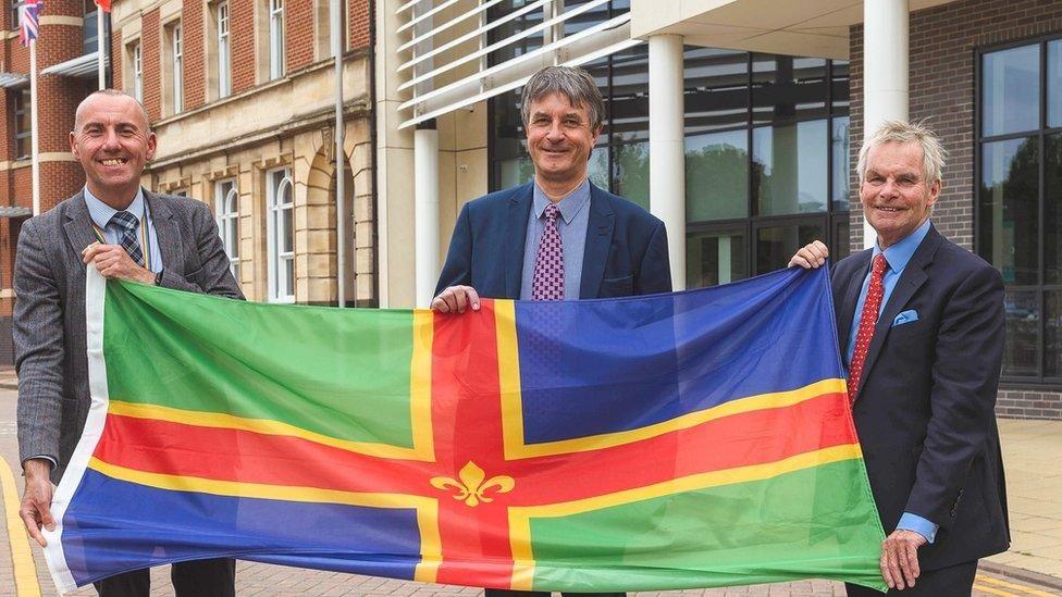 Three men wearing business suits stand holding a large Lincolnshire flag. The flag is of green and blue quarters divided by a red and yellow cross with a fleur-de-lys in the middle.