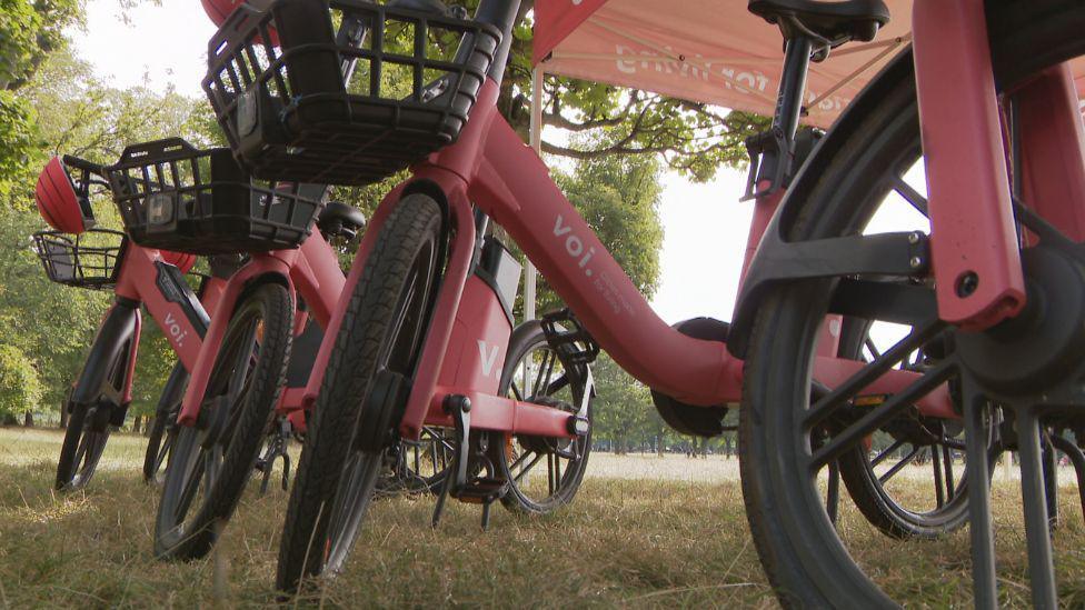 A row of red bikes with black wheels and black baskets on the front. They have the name Voi in white letters on the frame.