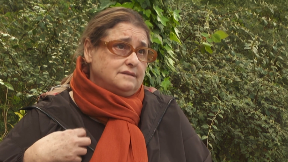 A woman in brown glasses, a brown coat and orange scarf looks to the side of the camera, stood in front of a hedge