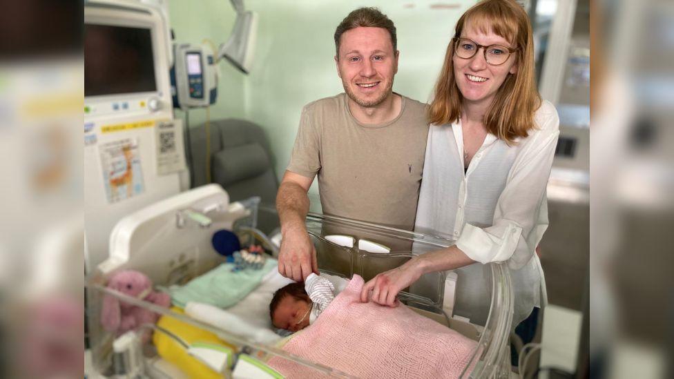 Pete Grimwade standing with his wife Charlotte in hospital- with baby Isabella wrapped in a pink blanket in an incubator