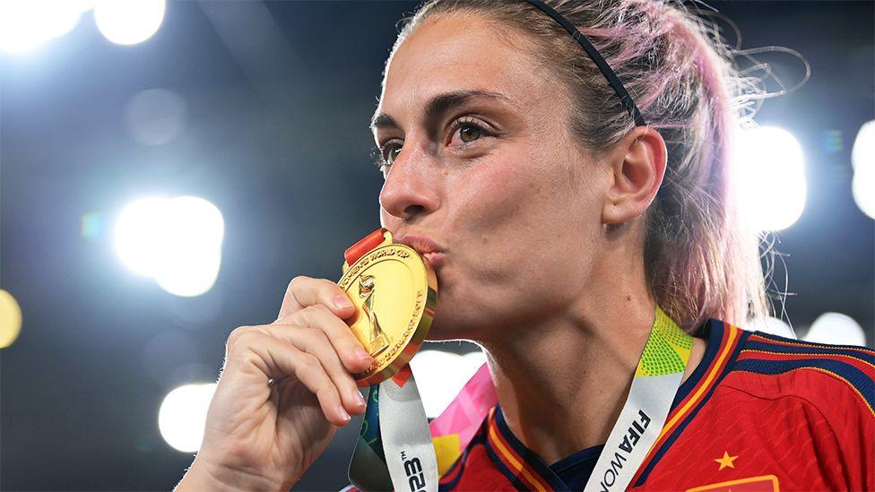 Alexia Putellas, wearing a red Spain football jersey, kissing a gold medal.