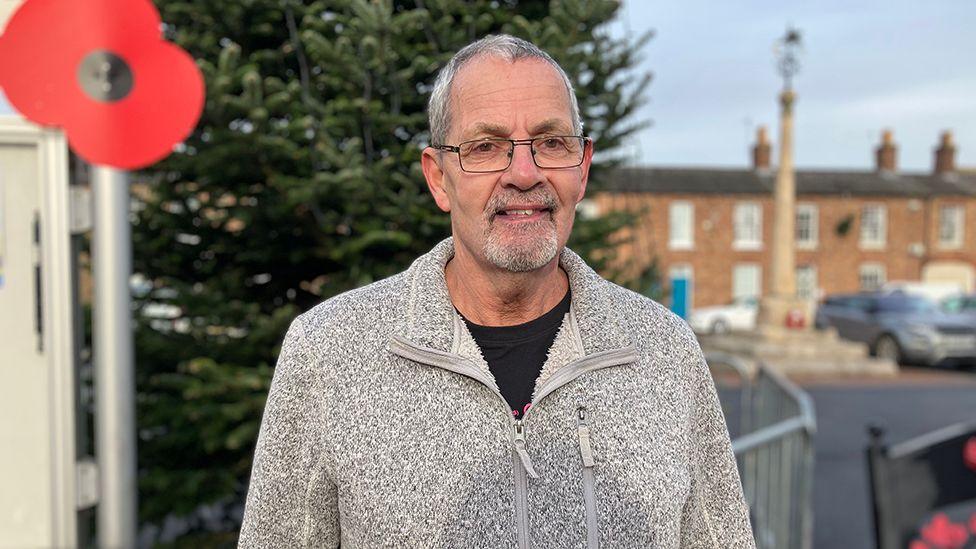 Wainfleet mayor councillor Steve Mitchell dressed in a grey top and wearing glasses standing in front of the Wainfleet Christmas tree