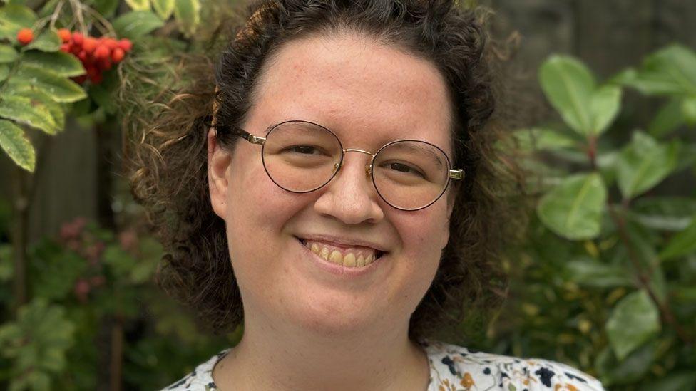 Emily Rhodes, with short curling dark hair haloed around her head, and round metal framed glasses, smiling at the camera with green shrubs with red berries behind her