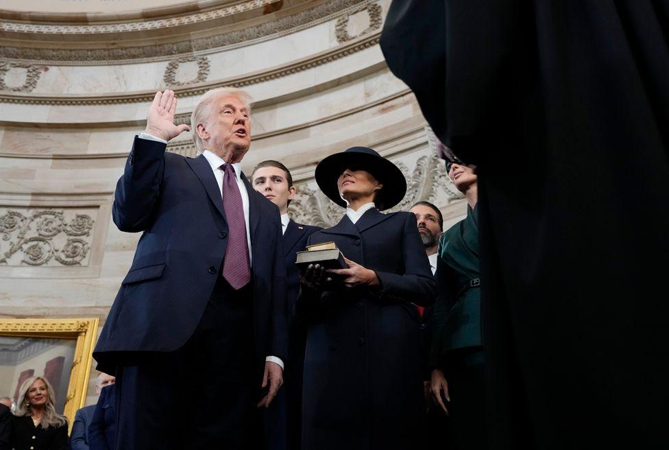 Donald Trump, wearing a dark suit and purple tie, stands with his right hand raised as Melania Trump, in a dark suit and dark hat, holds the Bible during the 60th Presidential Inauguration.  The curve of the Rotunda ceiling is in the background 