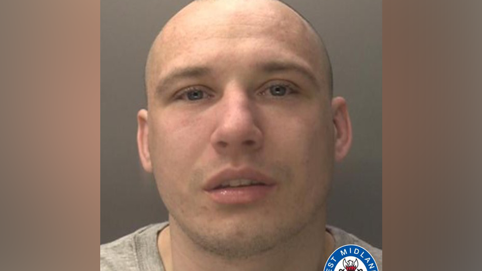 A police mug shot of a man with a shaved head looking at the camera. He is wearing a grey t-shirt. 