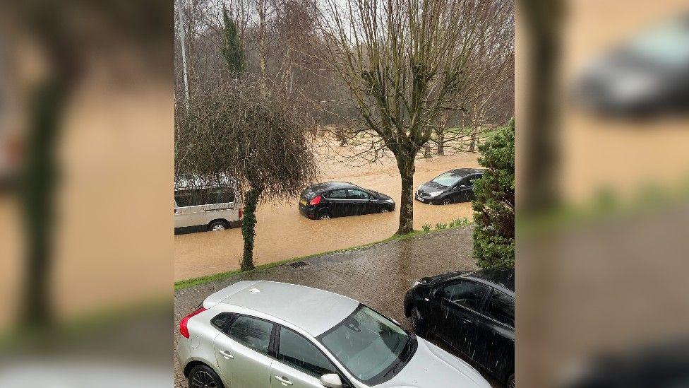 A flooded road seen from the upper window of a house, with cars submerged up the upper part of their wheels.