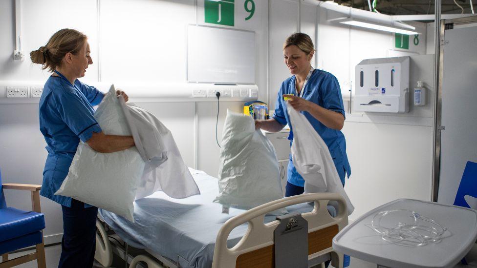 Two female nurses, both with blonde hair wearing blue scrubs, stand either side of a bed in a brightly-lit medical ward. They are putting pillow cases on pillows.