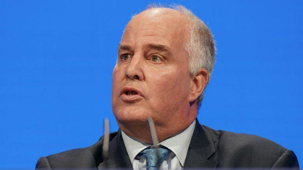 Andrew RT Davies speaking in front of a blue background into microphones. He is wearing a black suit, white shirt and blue tie.