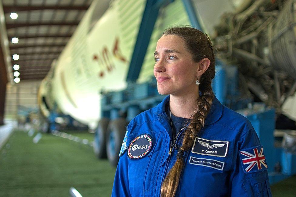 Rosemary Coogan standing in front of a Saturn V rocket, on its side, which was used for the Apollo missions. The rocket is defocussed and stretches off into the distance. Rosemary is dressed in her blue jumpsuit with her name on the right hand side, the European Space Agency logo on the left and the Union Jack on her left arm. Her brown hair is tied in a long plait over her left shoulder. She is wearing small dangly earrings with a small astronaut perched in the lap of a crescent moon.