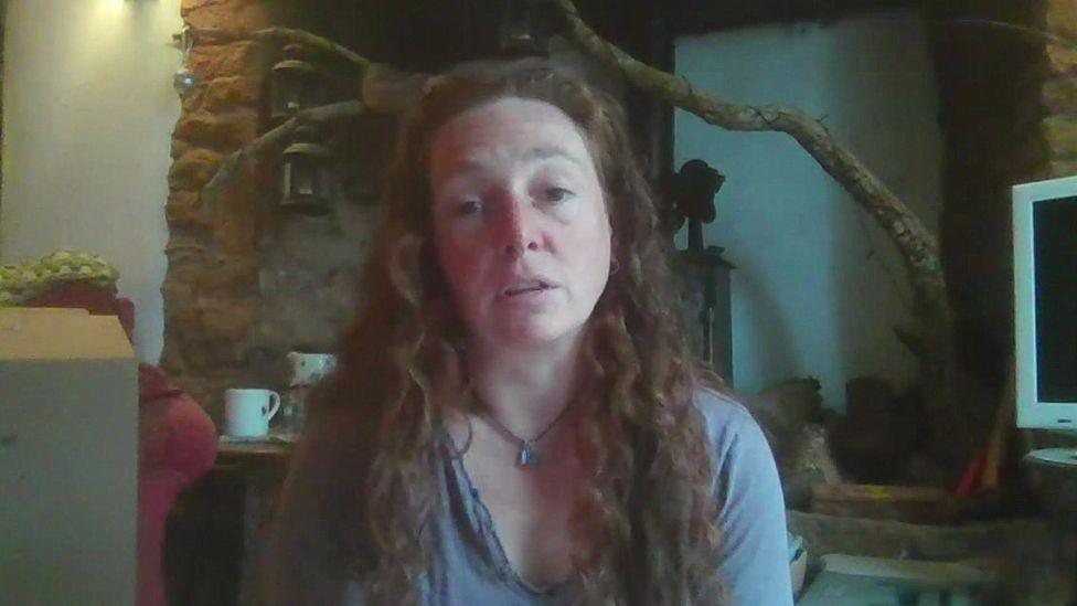 Emma Olliff, who has long, curly auburn hair, is wearing a lilac top and a necklace with a small pendant. She is sitting indoors in front of a stone fireplace.