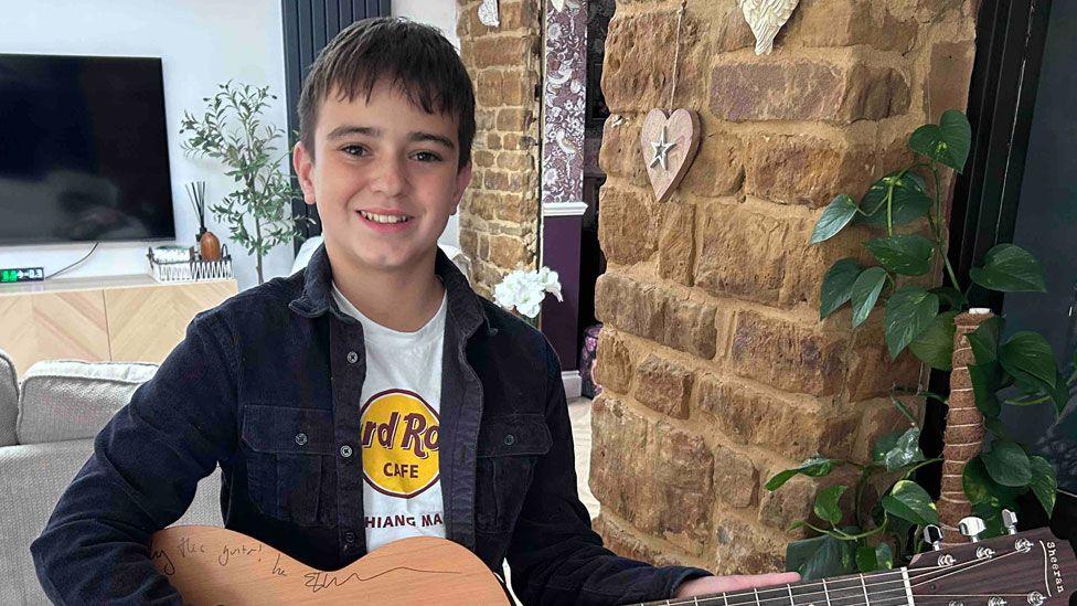 Jakob Wojcik, who has short black hair and is wearing a black open shirt over a white T-shirt. He is holding a guitar, which is signed along its top body edge, and is smiling. He is standing a TV behind him on the wall and a brick wall to his right. 
