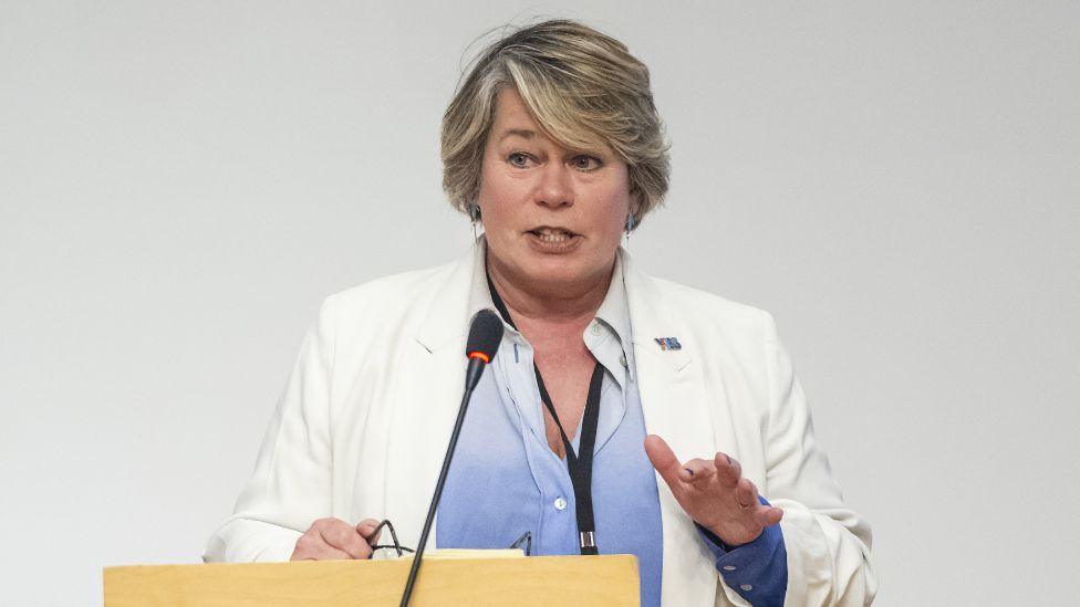 A woman with blonde hair, wearing a white suit and blue shirt, speaks at a podium in front of a grey background 