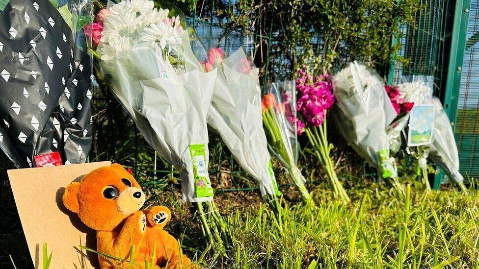 A brown stuffed soft toy bear in from of an envelope and bunches of flowers, some pink and some white, on grass next to a fence at the scene of a fatal accident