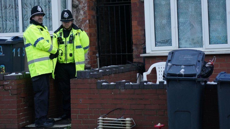 Two police officers in uniform standing outside a property with bins in front of it