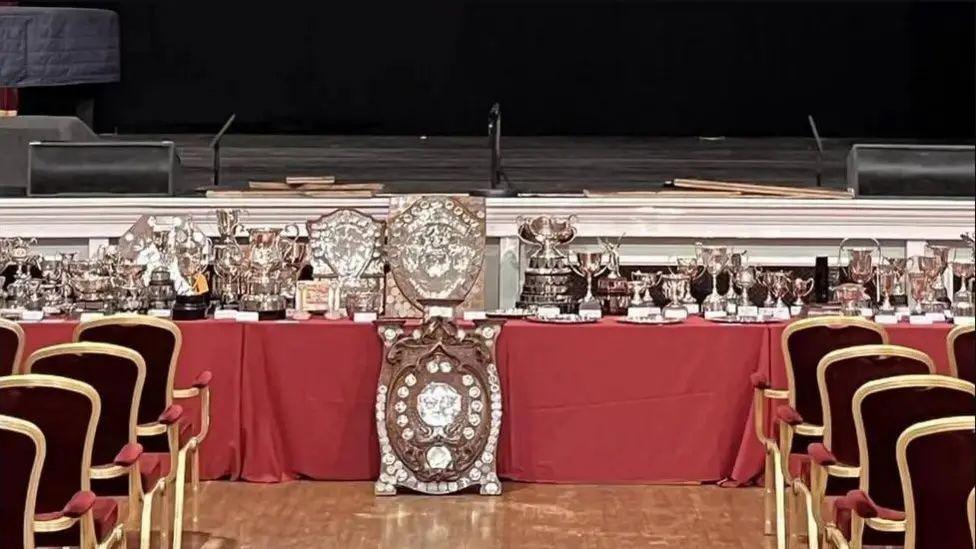 A table of trophies, with a large shield in the foreground and chairs on either side, there is a stage in the background.