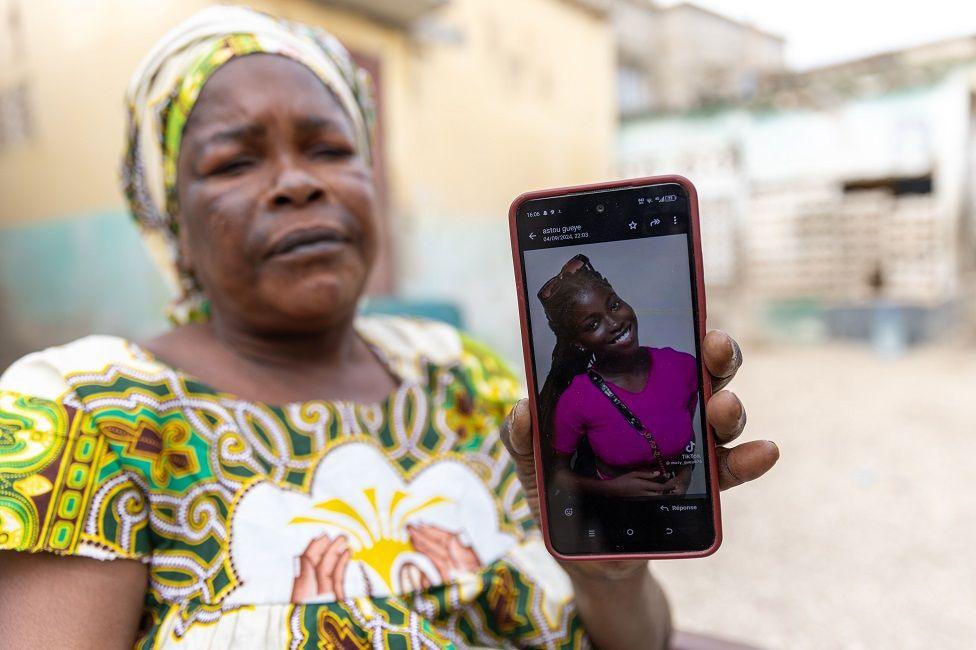 A grieving woman holds up a smartphone, showing a photo of her daughter smiling and dressed in pink.