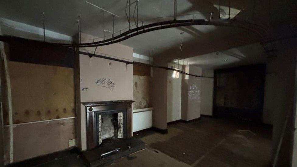 The railway track is in place in the ceiling but the walls have graffiti on them and windows are boarded up with debris and damp visible.