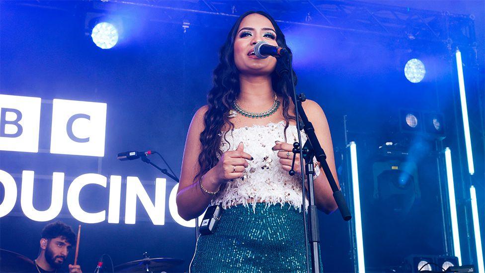 A woman on stage wearing a white top, singing into a black microhone. Behind her is a sign lit up in white which reads BBC Introducing, with a man in front of the sign playing drums.