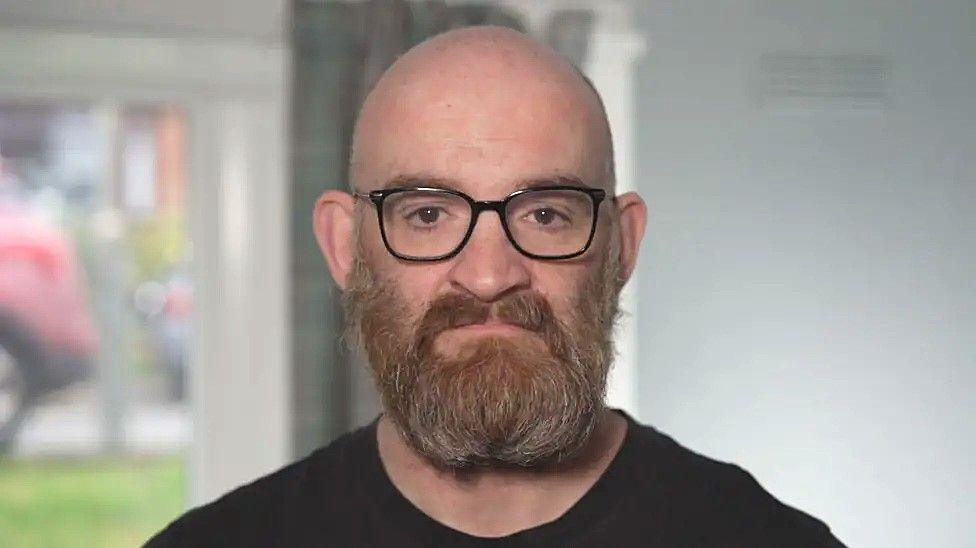 Keith Macaldowie, a bald man with a brown/red/grey beard looks sternly into the camera. He wears glasses and a black t-shirt.