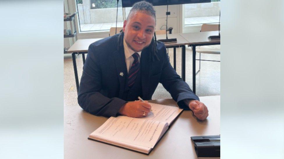 Stephen Lewis Elms smiling at the camera and signing a book. He is wearing a suit and a tie.