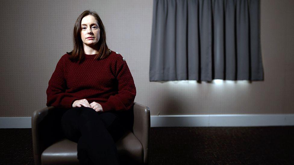 Business coach Rebecca Amin sits on a leather chair. She is wearing a maroon sweater with buttons on the shoulder. Behind her there is a window with curtains pulled shut.
