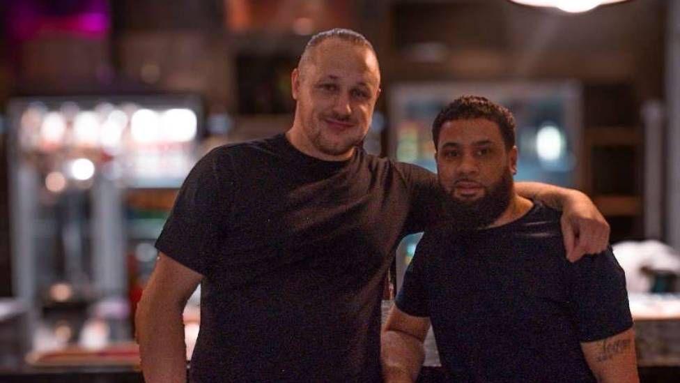 Two men in black t-shirts. On the left a man has his hair tied back, the man on the right has dark hair and a thick, dark beard