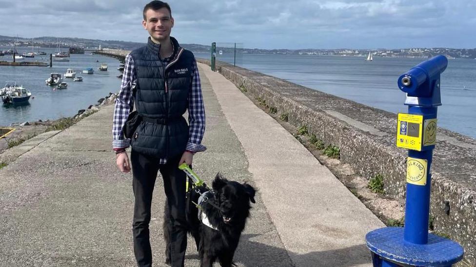 A young man stands on a coastal pathway beside a black guide dog, holding its harness. Boats are docked in the sea to the left, with land visible across the water. A blue public telescope is mounted on a concrete base to the right. The scene is calm and partly cloudy.