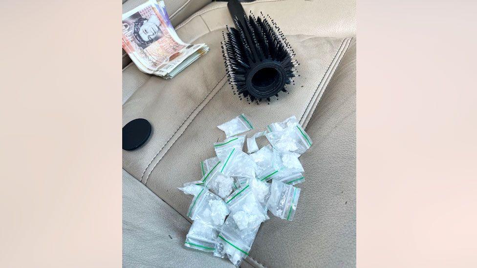 A round hairbrush with its top off laying on a cream leather car seat, next to a number of small plastic clear packets and a bundle of cash notes.