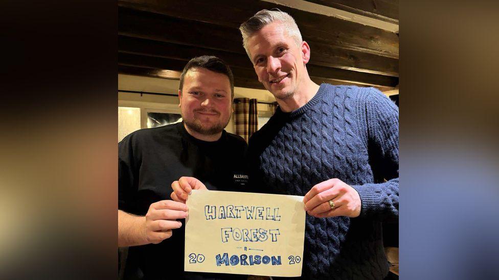 Jay Gurney and Steve Morison pose inside a pub and hold a piece of A4 paper with the words 'Hartwell Forest - Morison' written crudely in blue ink.