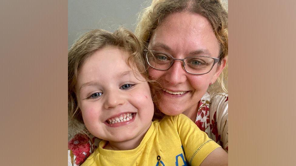 Athena Rowley cuddling her son Robert as they take a selfie together. She is wearing glasses and has curly hair while he wears a yellow t-shirt.